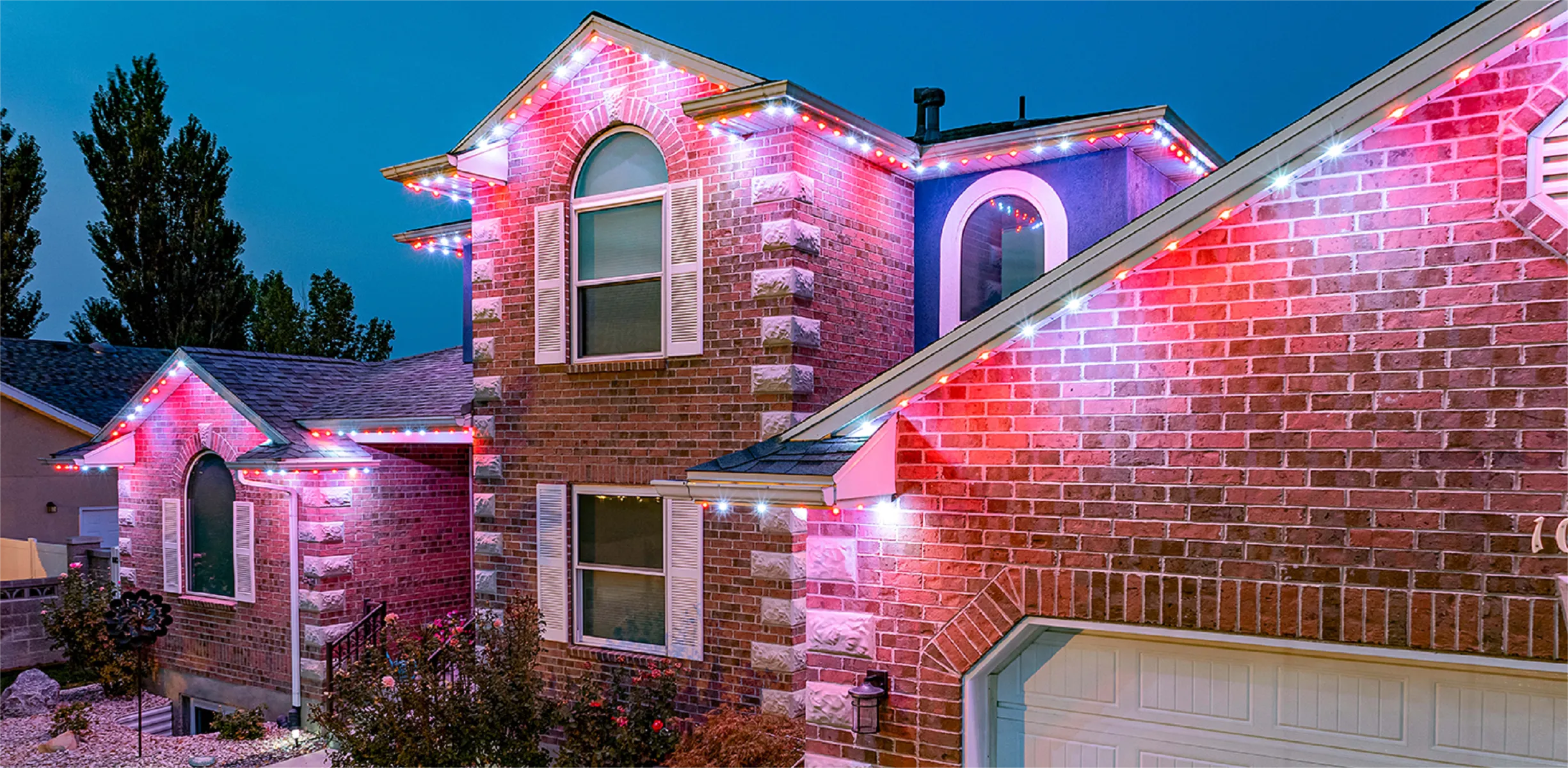 Two-story brick home at dusk with pink and white permanent lighting along every roofline